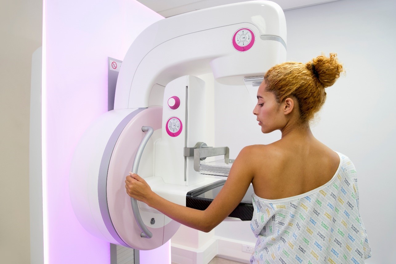 Woman standing beside a white mammogram machine with her robe pulled off one shoulder for the procedure
