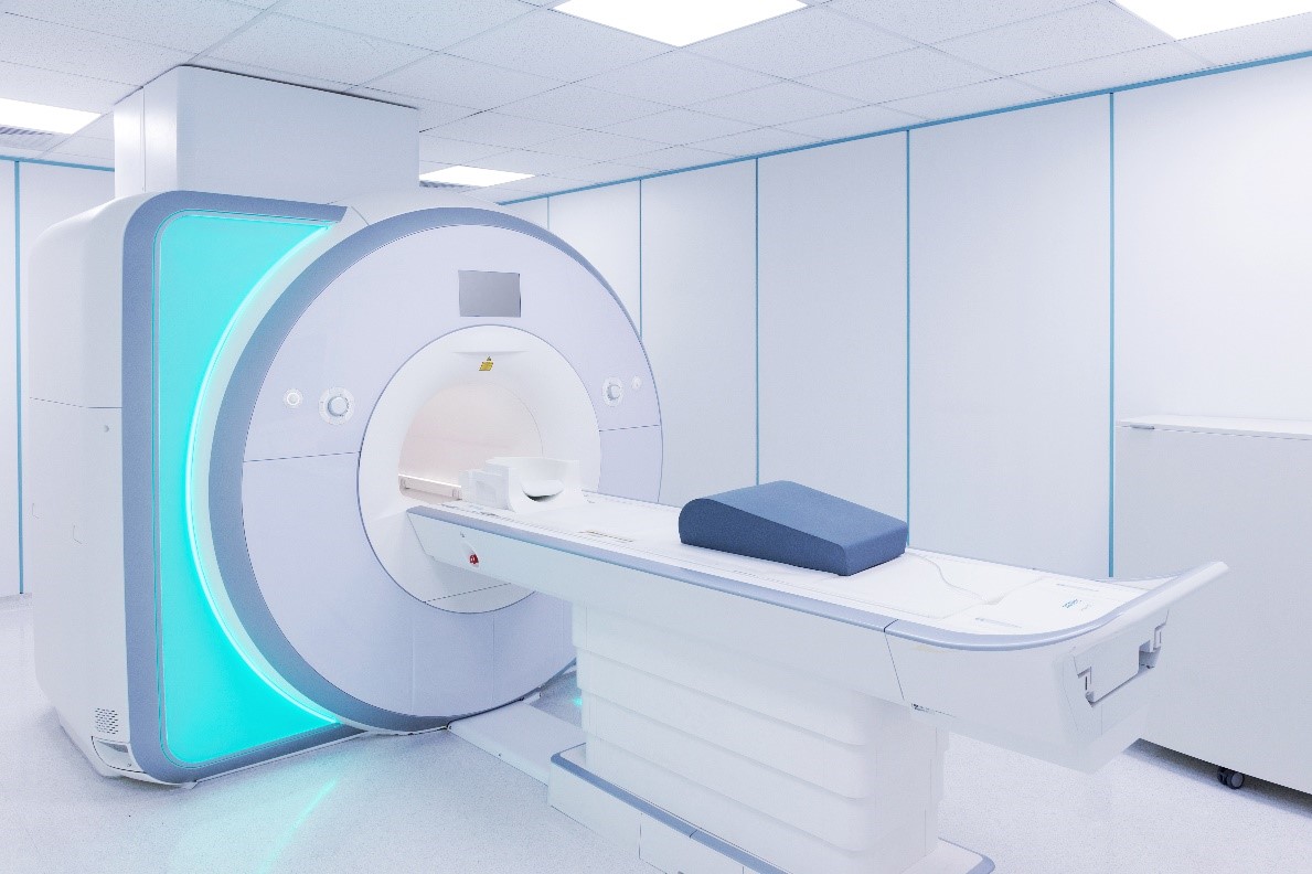 A white MRI machine in a brightly lit white room with a blue cushion on the machine’s bed